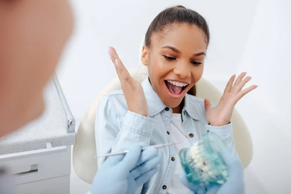 Foco seletivo da mulher americana africana excitada em aparelho gestual perto dentista segurando dentes modelo — Fotografia de Stock