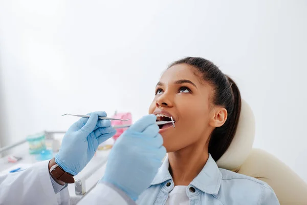 Стоматолог в латексных перчатках держит стоматологическое оборудование рядом с африканским американским пациентом с открытым ртом и брекетами — стоковое фото