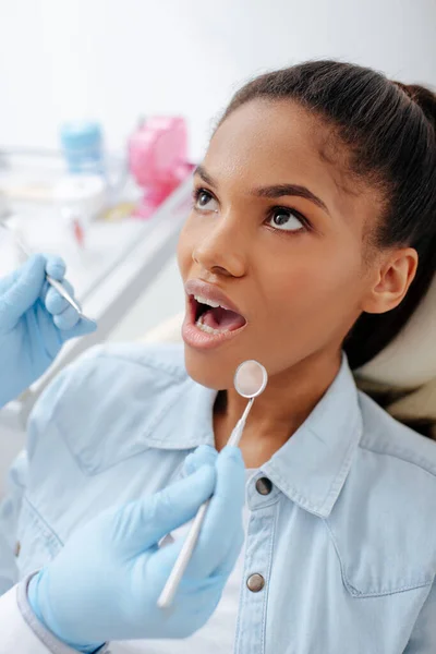 Dentista in guanti di lattice in possesso di attrezzature dentali vicino al paziente afroamericano con bocca aperta — Foto stock