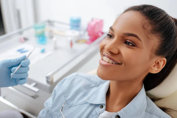 Orthodontiste dans la tenue des instruments dentaires près heureux patient afro-américain dans les appareils dentaires — Photo de stock