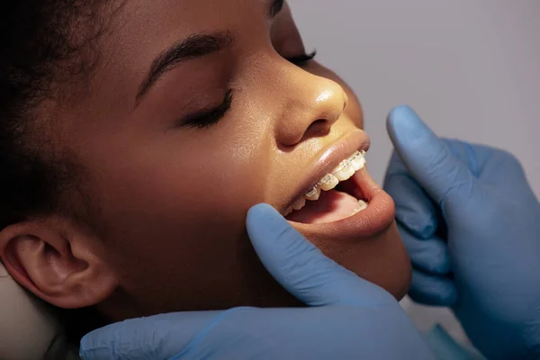 Orthodontiste en gants de latex près heureuse femme afro-américaine en bretelles — Photo de stock