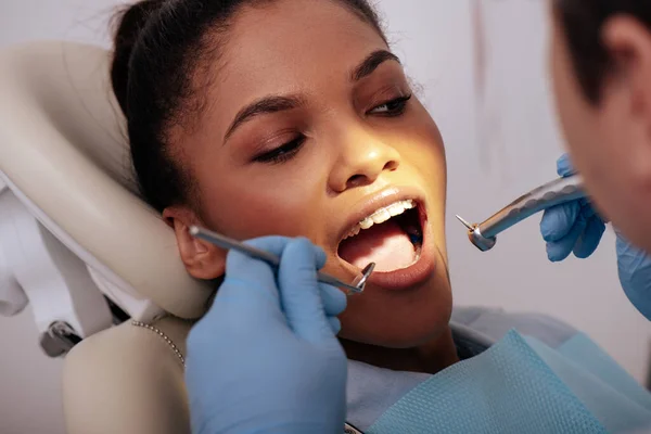 Foco seletivo do dentista em luvas de látex segurando instrumentos dentários perto atraente mulher afro-americana em aparelho — Fotografia de Stock