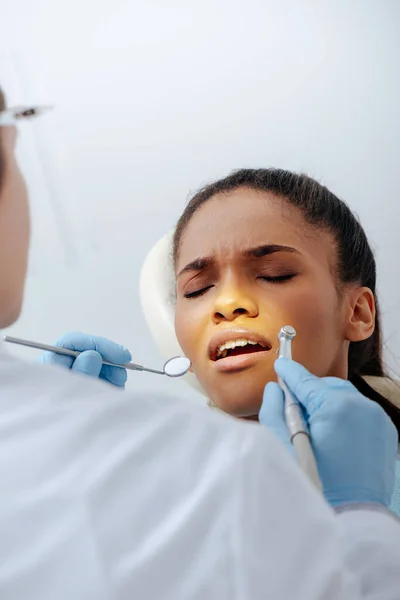 Enfoque selectivo de ortodoncista en guantes de látex que sostienen instrumentos dentales cerca de la mujer afroamericana molesta en frenos - foto de stock