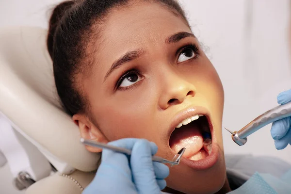 Вибірковий фокус стоматолога в латексних рукавичках, що тримає зубні інструменти біля привабливої афроамериканської жінки з відкритим ротом — стокове фото