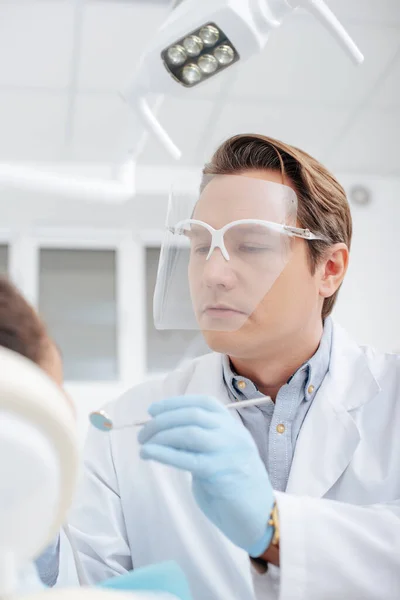 Селективное внимание стоматолога в защитном щите лица, держащего зубное зеркало рядом с африканской американкой — стоковое фото