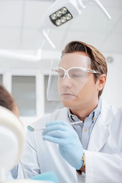 Messa a fuoco selettiva del dentista in protettivo viso scudo tenendo specchio dentale vicino donna afro-americana — Foto stock