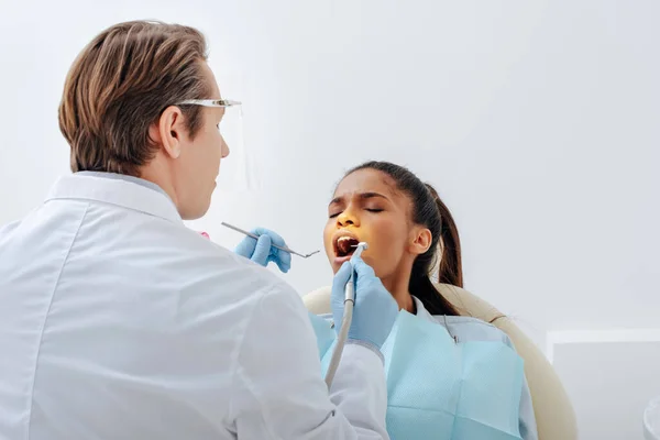 Dentiste en gants de latex et écran facial tenant des instruments dentaires près de femme afro-américaine attrayante avec bouche ouverte — Photo de stock
