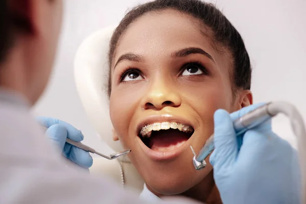 Foyer sélectif du dentiste dans des gants en latex tenant des instruments dentaires près heureuse femme afro-américaine avec bouche ouverte — Photo de stock