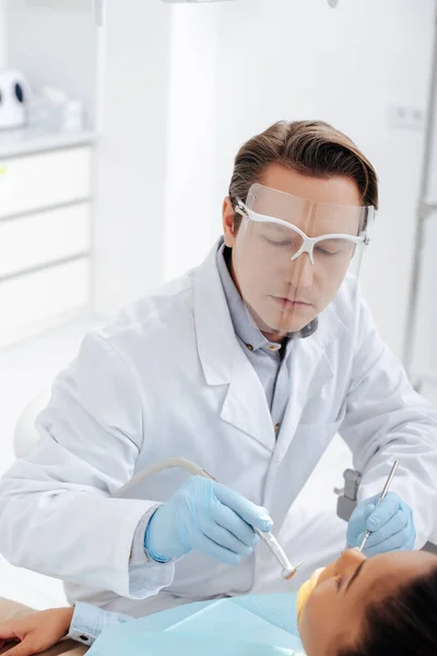 Foyer sélectif du dentiste dans le bouclier facial et gants en latex tenant des instruments dentaires près de la femme afro-américaine — Photo de stock
