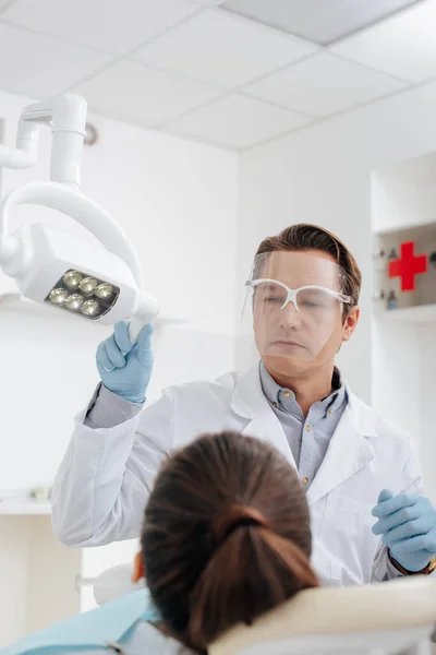 Enfoque selectivo del dentista en el escudo facial y guantes de látex tocando lámpara médica cerca del paciente - foto de stock