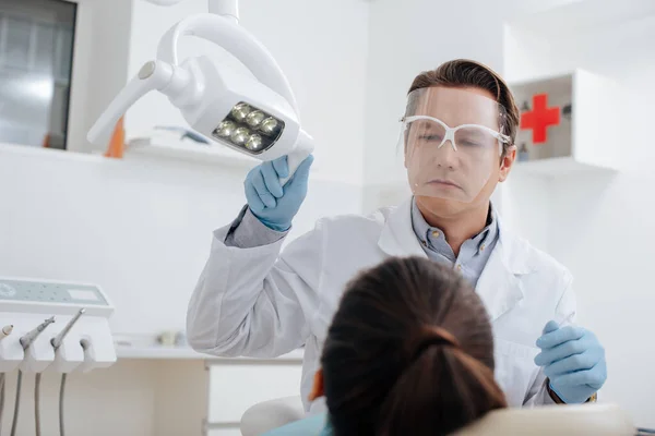 Enfoque selectivo del dentista en el escudo facial y guantes de látex tocando lámpara médica cerca de la mujer - foto de stock