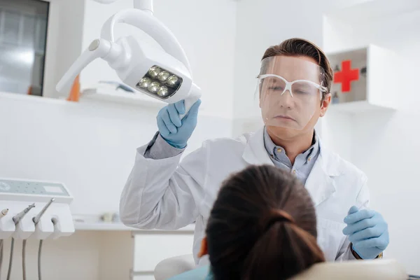 Selective focus of dentist in face shield and latex gloves touching medical lamp near woman — Stock Photo