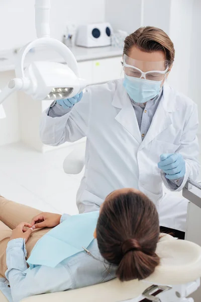 Messa a fuoco selettiva del dentista in faccia scudo, maschera medica e guanti in lattice toccare lampada medica vicino donna afro-americana — Foto stock