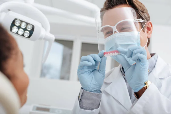 Селективный фокус стоматолога в медицинской маске, защитных и латексных перчатках, держащих зубной протез рядом с африканской американкой — стоковое фото