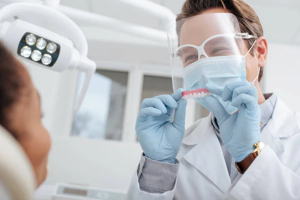 Foco seletivo do dentista em máscara médica, protetor facial e luvas de látex segurando prótese dentária perto de mulher afro-americana — Fotografia de Stock