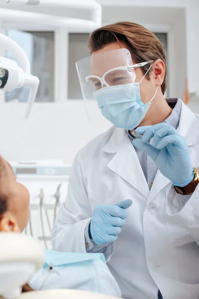 Селективный фокус стоматолога в медицинской маске, защитных и латексных перчатках, держащих стоматологический инструмент рядом с африканской американкой — стоковое фото