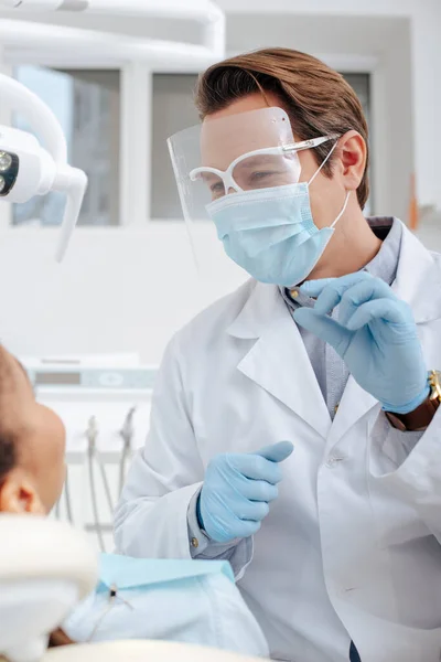 Foyer sélectif du dentiste dans le masque médical, écran facial et gants en latex tenant instrument dentaire près de la femme afro-américaine — Photo de stock