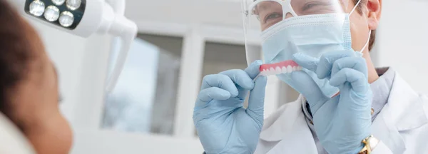 Prise de vue panoramique du dentiste en masque médical, écran facial et gants en latex tenant une prothèse dentaire près d'une femme afro-américaine — Photo de stock