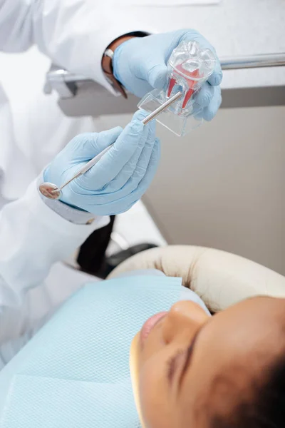 Messa a fuoco selettiva del dentista in possesso di strumento dentale modello dente pulito e paziente afro-americano — Foto stock