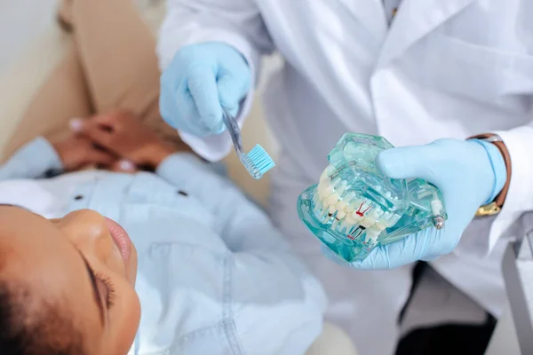 Enfoque selectivo del dentista en guantes de látex que sostienen el modelo de dientes y el cepillo de dientes cerca del paciente afroamericano - foto de stock