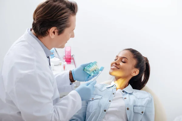 Foyer sélectif du dentiste dans des gants en latex tenant modèle de dents et brosse à dents près heureux patient afro-américain — Photo de stock