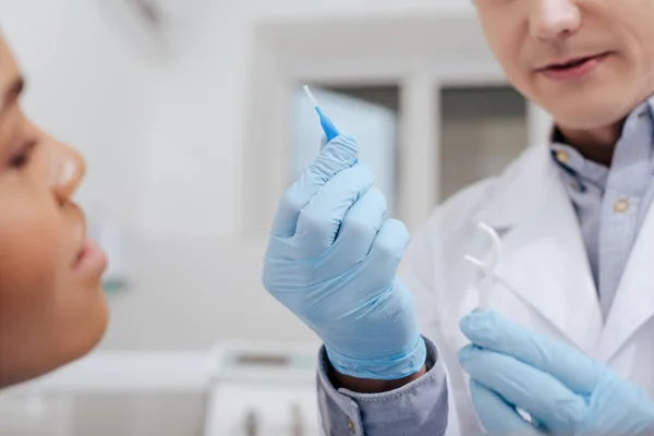 Vue recadrée du dentiste tenant un bâton de fil dentaire et une brosse de nettoyage inter dentaire près d'une femme afro-américaine — Photo de stock