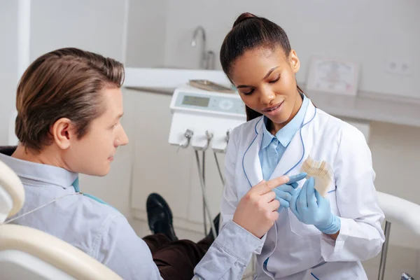 Hombre señalando con el dedo a la paleta de dientes cerca del dentista afroamericano - foto de stock