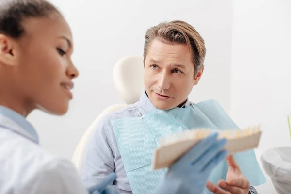 Foco seletivo do paciente olhando para dentista afro-americano segurando paleta de dentes — Fotografia de Stock