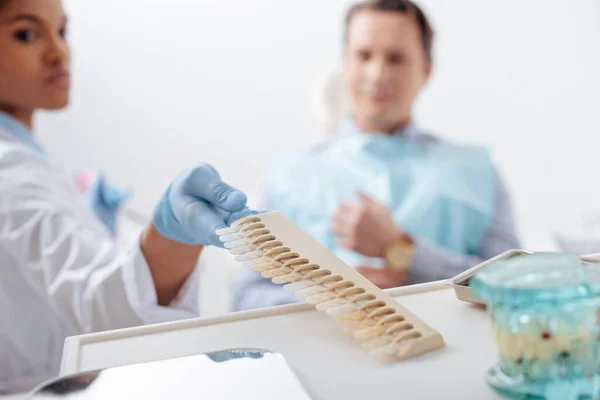 Focalizzazione selettiva del dentista afroamericano in guanto di lattice che prende la tavolozza dei denti vicino al paziente — Foto stock
