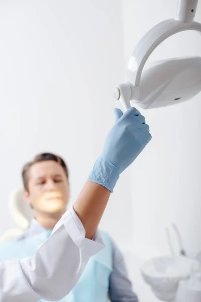 Селективный фокус африканского американского стоматолога в латексной перчатке касающийся медицинской лампы рядом с пациентом — стоковое фото