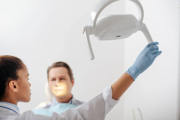 Selektiver Fokus attraktiver afrikanisch-amerikanischer Zahnarzt in Latexhandschuhen, die medizinische Lampe in der Nähe des Patienten berühren — Stockfoto