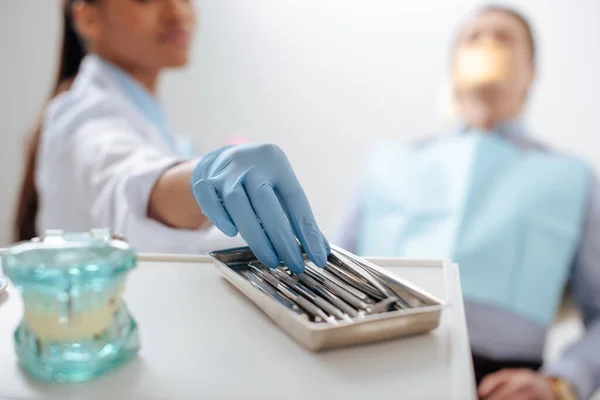 Selective focus of african american dentist in latex glove taking dental instrument near patient — Stock Photo