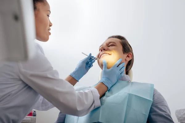 Селективное внимание африканского американского стоматолога в латексных перчатках, держащего стоматологический инструмент рядом с счастливым пациентом — стоковое фото