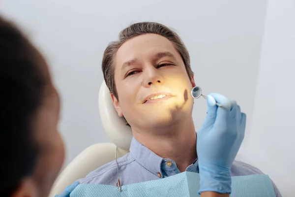 Foco seletivo do dentista afro-americano em luva de látex segurando espelho dental perto de paciente bonito — Fotografia de Stock
