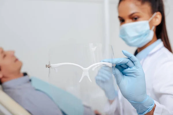 Foyer sélectif du dentiste afro-américain dans le masque médical tenant le bouclier facial près du patient — Photo de stock