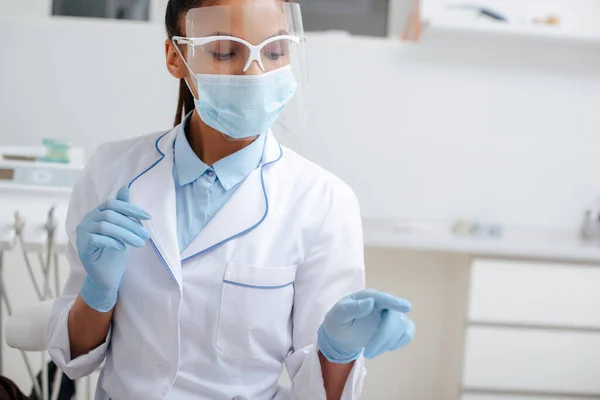 Afrikanisch-amerikanischer Zahnarzt mit medizinischer Maske, Gesichtsschutz und Latexhandschuhen, der mit dem Finger in die Klinik zeigt — Stockfoto