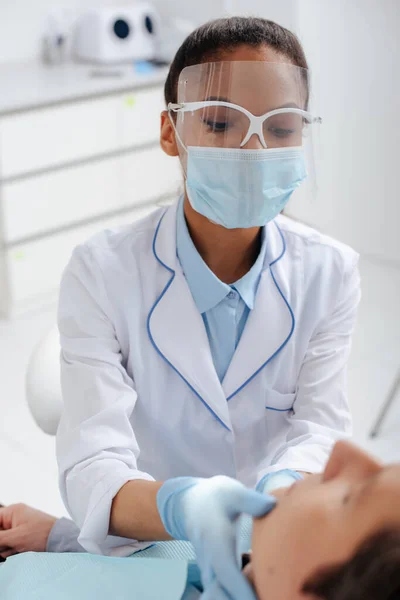 Messa a fuoco selettiva del dentista afroamericano in maschera medica e scudo facciale toccare il paziente — Foto stock