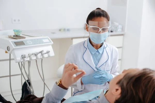 Foco seletivo do dentista afro-americano em máscara médica e protetor facial perto do paciente na clínica — Fotografia de Stock