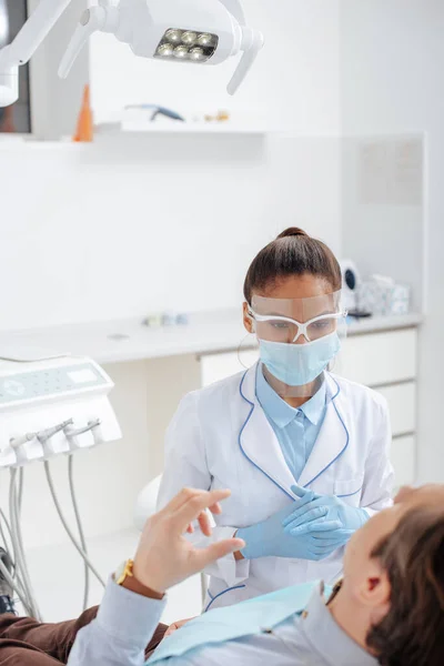 Селективный фокус африканского американского стоматолога в медицинской маске и щите лица рядом с мужчиной в клинике — стоковое фото