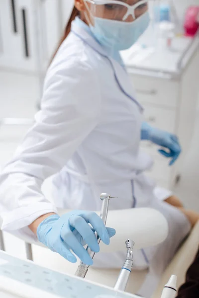 Enfoque selectivo del dentista afroamericano en máscara médica y escudo facial tomando instrumento dental inoxidable - foto de stock