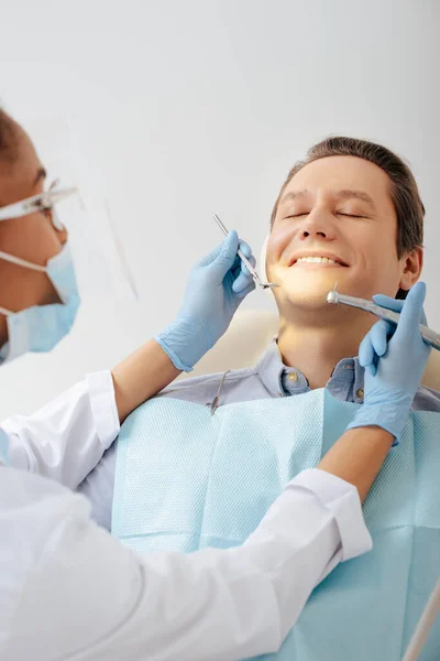 Foco seletivo do dentista afro-americano em máscara médica e protetor facial segurando instrumentos dentários perto do paciente feliz — Fotografia de Stock