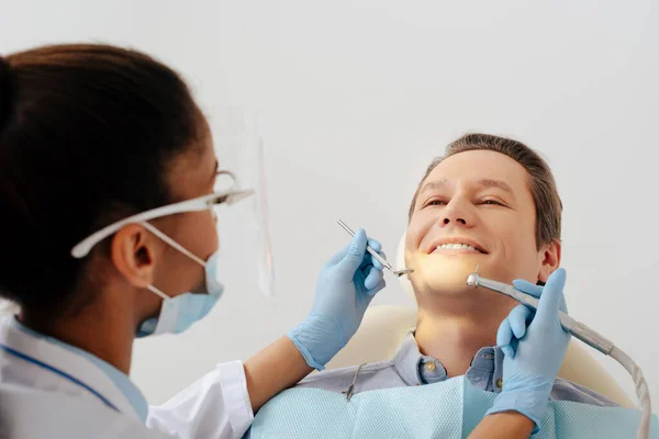 Foyer sélectif du dentiste afro-américain dans le masque médical et le bouclier facial tenant les instruments dentaires près du patient joyeux — Photo de stock