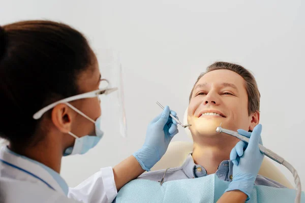 Selective focus of african american dentist in medical mask and face shield holding dental instruments near cheerful patient — Stock Photo