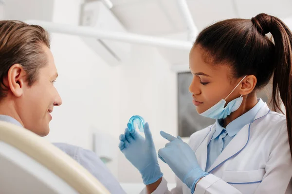 Foyer sélectif du dentiste afro-américain dans des gants en latex pointant du doigt le dispositif de retenue près du patient heureux — Photo de stock