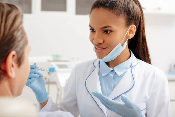 Вибірковий фокус щасливого афроамериканського стоматолога в латексних рукавичках, що вказують руками на зубну щітку біля пацієнта — стокове фото