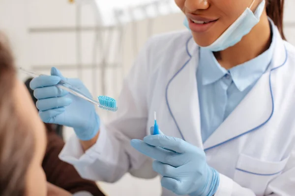 Vista recortada del dentista afroamericano en guantes de látex con cepillo de dientes y cepillo de limpieza interdental cerca del paciente - foto de stock