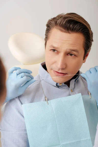Selektiver Fokus des Zahnarztes in Latexhandschuhen, die Zahnseide in der Nähe eines hübschen Patienten halten — Stockfoto