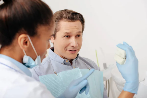 Messa a fuoco selettiva del dentista afroamericano in guanti di lattice che punta con il dito al modello di denti vicino al paziente — Foto stock