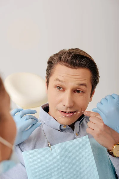 Селективный фокус африканского американского стоматолога в латексных перчатках, держащего зубную нить рядом с красивым пациентом, указывающим пальцем в лицо — стоковое фото