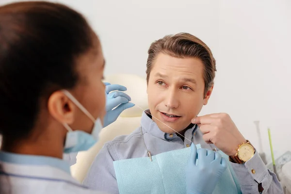 Selektiver Fokus eines afrikanisch-amerikanischen Zahnarztes in Latexhandschuhen, der Zahnseide in der Nähe des Patienten hält und mit dem Finger auf das Gesicht zeigt — Stockfoto